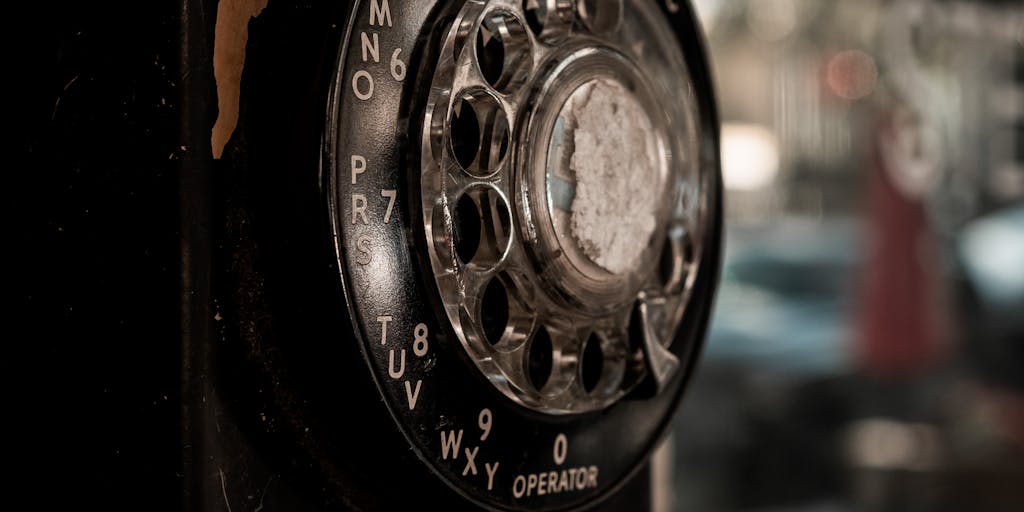 A close-up shot of a vintage rotary phone, with the dial partially turned to '917'. The phone is placed on a weathered wooden surface, perhaps a park bench or an old desk, with a blurred cityscape of New York City in the background, subtly hinting at the area code's location. The lighting should be warm and nostalgic, emphasizing the retro feel of the phone and the connection to a bygone era of communication in the city.