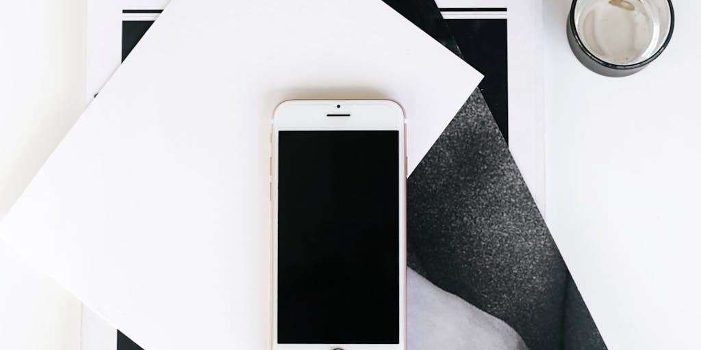 An artistic flat lay of a smartphone displaying the Pinterest app, surrounded by various marketing materials such as brochures, business cards, and a cup of coffee. The background is a textured wooden table, and the composition highlights the integration of digital marketing with traditional methods.