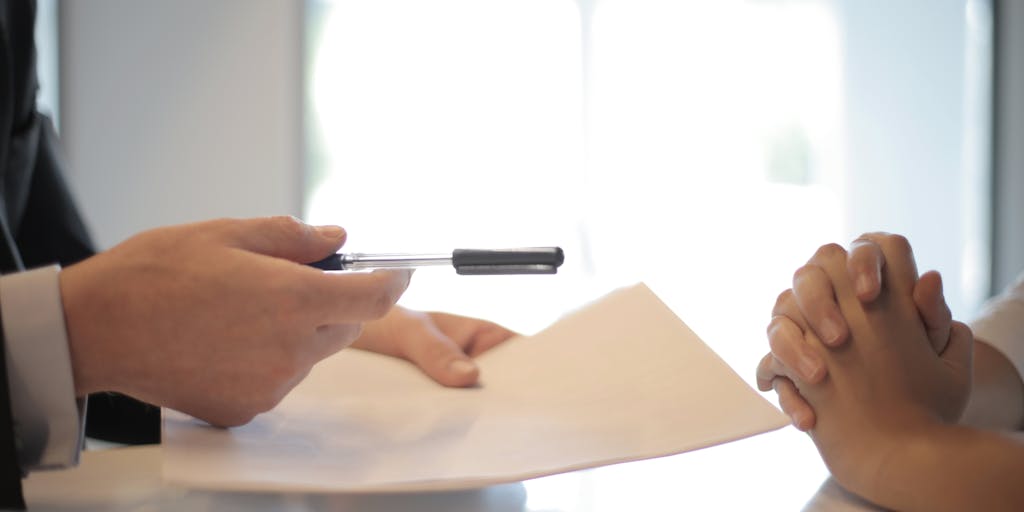 A close-up shot of a business professional's hands signing a legal agreement on a wooden desk, with a pen in hand and a focused expression. In the background, a blurred image of a laptop displaying a digital contract, symbolizing the importance of legal agreements in safeguarding business interests.