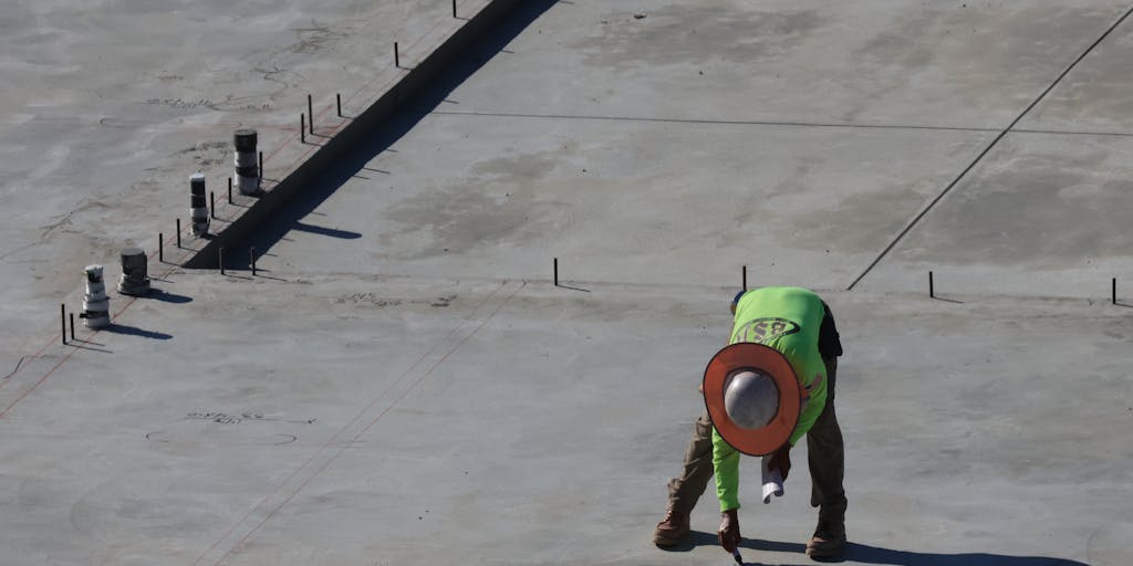 A close-up shot of a hard hat with a visible crack, resting on a construction site floor scattered with tools and debris. In the background, a blurred image of a construction worker sitting on a bench, holding their injured arm, symbolizing the aftermath of an accident.