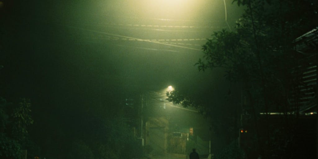 A shadowy figure standing next to a deserted road at night, looking back over their shoulder with a worried expression. The scene is illuminated by the faint glow of a streetlight, creating an eerie atmosphere. The background features blurred headlights of a car approaching, symbolizing the impending consequences of their actions.