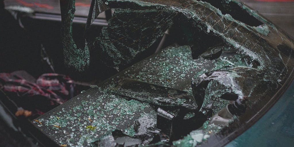 A close-up shot of a crumpled car bumper with paint scratches and debris scattered around, suggesting a recent accident. In the background, a blurred police car with flashing lights is visible, emphasizing the urgency of the situation. The focus is on the damage, highlighting the aftermath of a hit and run.