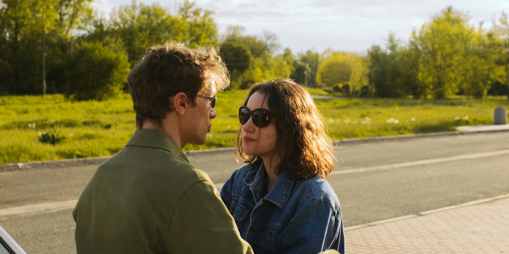 A close-up shot of a person looking nervously over their shoulder while sitting in a café, with a blurred police car visible through the window. The expression on their face conveys anxiety and suspicion, hinting at the idea of being watched.