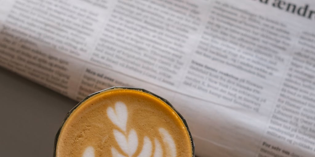 A cozy morning scene featuring a person sipping coffee while reading a newspaper, with sunlight streaming through the window. The table is cluttered with everyday items like a half-eaten croissant, a pair of glasses, and a potted plant, capturing the essence of routine and comfort.