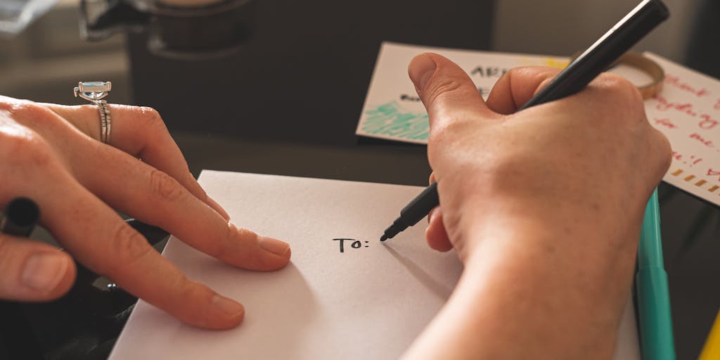 A close-up shot of a hand writing an address on a single line using a colorful gel pen on a textured piece of stationery. The background is softly blurred to emphasize the writing process, with a ruler and a coffee cup in the frame to add a cozy, creative workspace vibe.