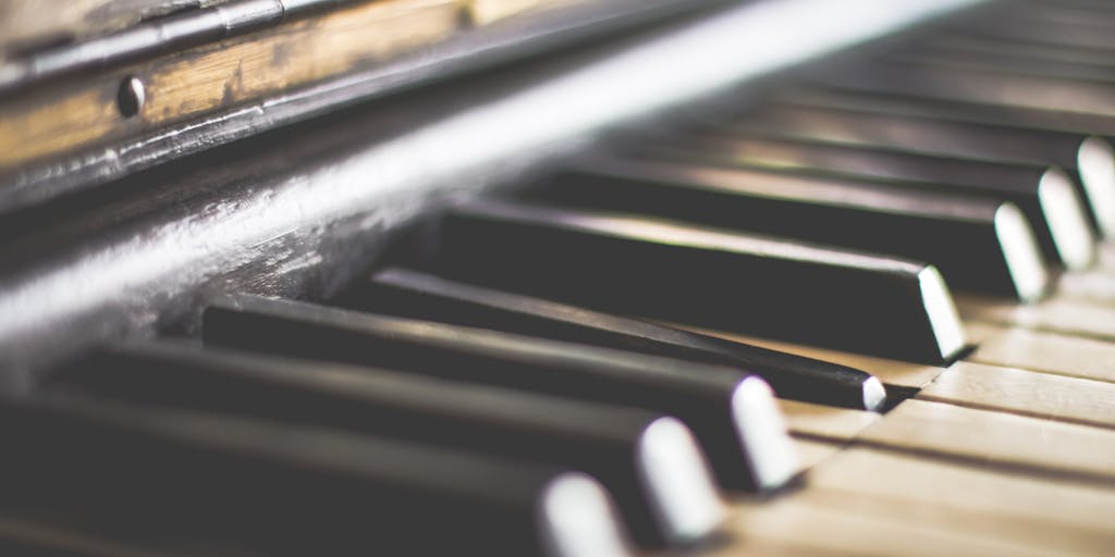 A close-up shot of a musician's hand playing a C note on a grand piano, with the focus on the keys and the hand's movement. The background is softly blurred to emphasize the elegance of the music being played.