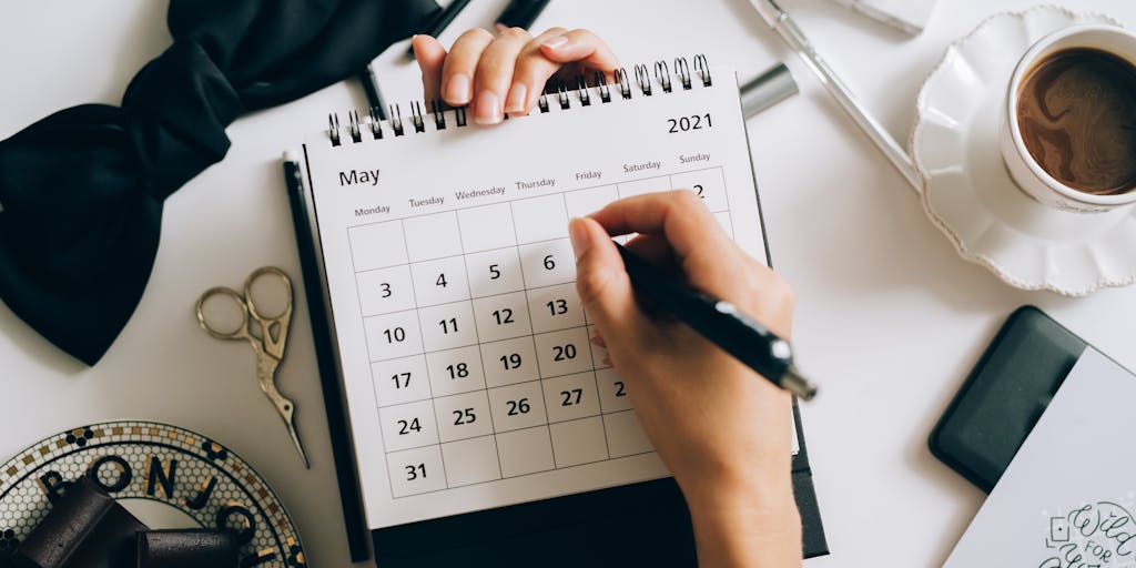 A close-up shot of a calendar page with the dates circled in red, showing a week and a half. The background features a desk with a laptop, coffee cup, and a clock indicating the time, symbolizing the passage of business days.