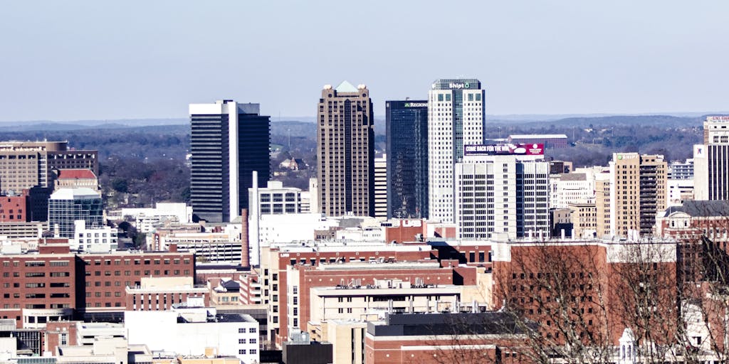 A dramatic shot of a high-rise office building in Birmingham, Alabama, with a lawyer in a sharp suit standing confidently in front of it, holding a briefcase. The skyline reflects the ambition and success of the highest-paid lawyers in the state.