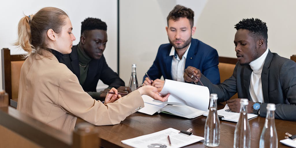 A group of diverse lawyers in formal attire gathered around a conference table, engaged in a discussion. The setting is a modern law office with large windows showcasing the Alaskan landscape, highlighting collaboration among the highest paid legal professionals.