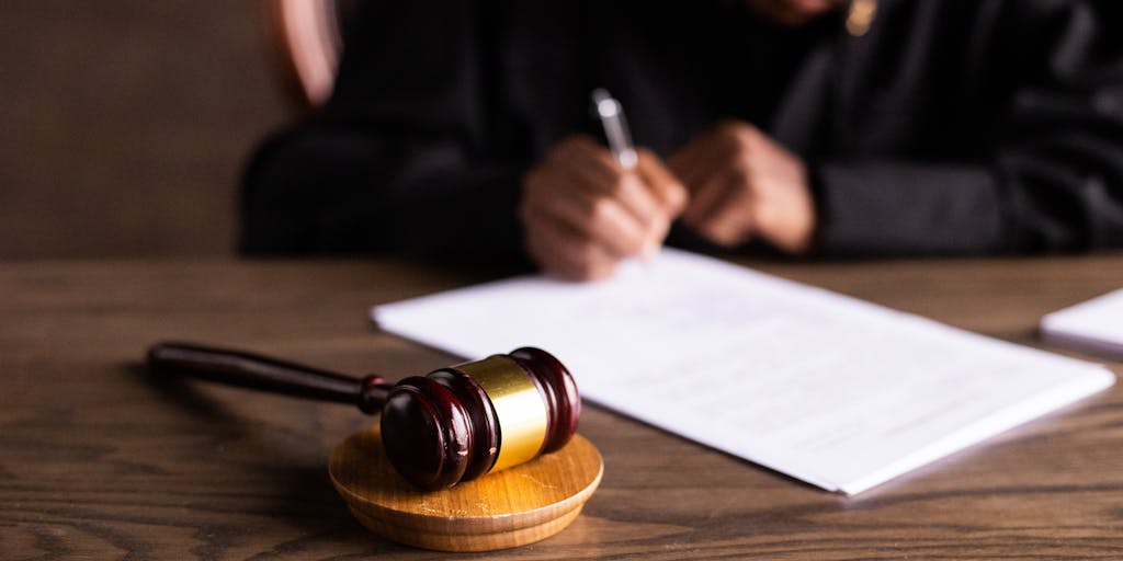 An artistic shot of a gavel and a stack of legal documents on a polished mahogany desk, with a blurred image of a courtroom in the background. The lighting is warm, creating a sense of authority and professionalism, highlighting the serious nature of legal work.