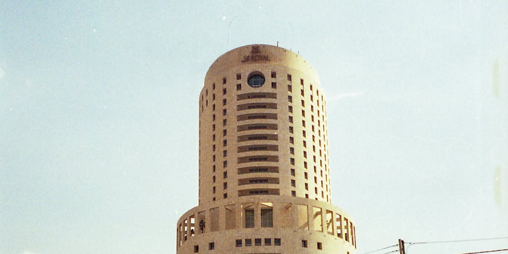 A dramatic shot of a high-rise office building in Amman, Jordan, with a lawyer in a tailored suit standing confidently in front of it, holding a briefcase. The skyline reflects the modernity and success associated with the highest-paid lawyers in the region.