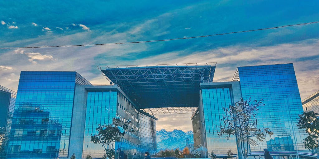 A dramatic portrait of a high-powered lawyer in a sleek, modern office overlooking the skyline of Almaty. The lawyer is dressed in an elegant suit, confidently reviewing legal documents with a cityscape backdrop at sunset, symbolizing success and ambition.