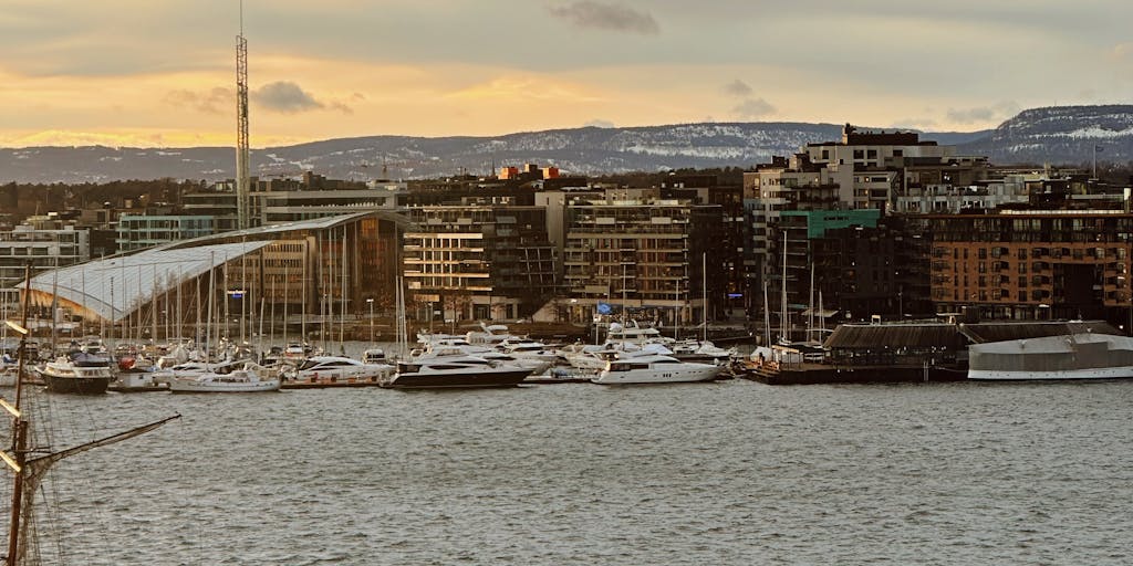 A dramatic shot of a high-rise office building in Oslo, reflecting the skyline at sunset. The building should have a modern architectural design, symbolizing the success and prestige of the highest paid lawyers in Norway. The warm colors of the sunset contrast with the cool glass facade, creating a visually striking image.