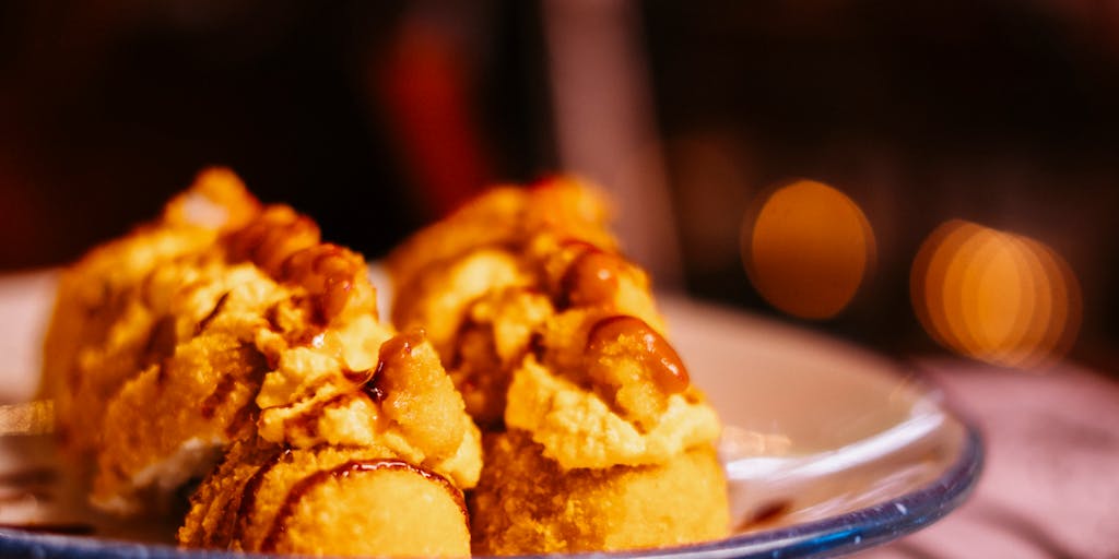 A close-up shot of a delicious, over-the-top dessert, such as a towering cake or a sundae overflowing with toppings. The dessert is placed on a vibrant tablecloth, and in the background, a group of friends can be seen gasping in awe, embodying the phrase 'Oh My Lanta!'