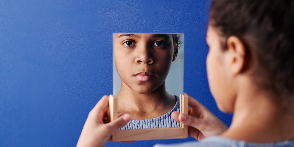 A creative portrait of a person standing in front of a large, ornate mirror. The subject is dressed in a vibrant outfit that contrasts with the muted colors of the room. The mirror reflects not only the subject but also the intricate details of the room, creating a layered composition that plays with the concept of reflection and identity.