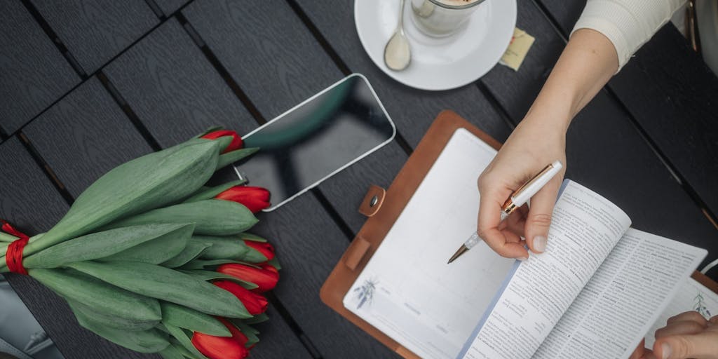 An open notebook on a wooden table, filled with handwritten notes and doodles. Surrounding the notebook are various items like a coffee cup, a smartphone, and a plant, showcasing a workspace that includes but is not limited to traditional study materials.