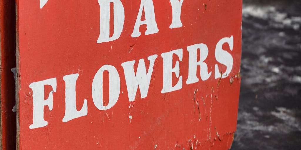 A close-up shot of a person holding a colorful, hand-painted sign that reads 'LLLP' in bold letters, surrounded by vibrant flowers and greenery. The background is softly blurred to emphasize the sign, creating a cheerful and inviting atmosphere.