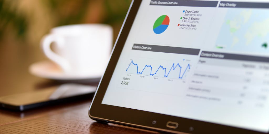 A close-up shot of a smartphone displaying the Dripify dashboard, with colorful graphs and metrics visible. The phone is placed on a wooden desk surrounded by coffee cups and notebooks, suggesting a productive work environment.