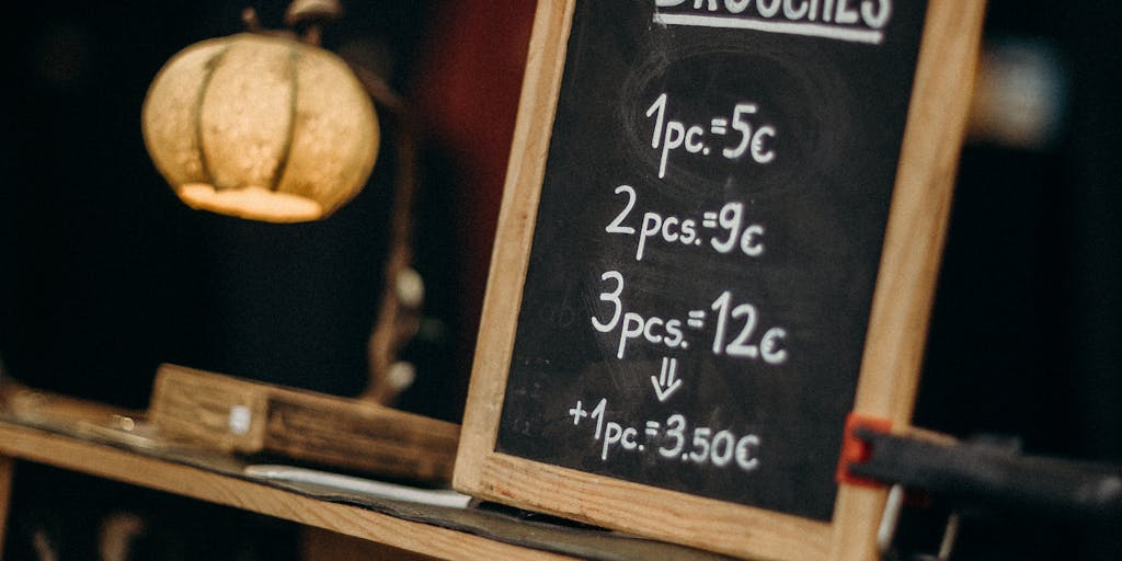 A close-up shot of a chalkboard menu displaying various Yamm pricing options, with colorful illustrations of dishes next to each price. The background features a cozy café setting with patrons enjoying their meals, creating a warm and inviting atmosphere.
