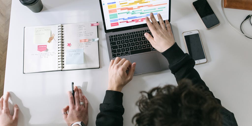 An overhead view of a desk cluttered with various pricing strategy documents, sticky notes, and a laptop open to the Alfred Pricing website. A cup of coffee sits beside the laptop, symbolizing the brainstorming process behind pricing decisions.