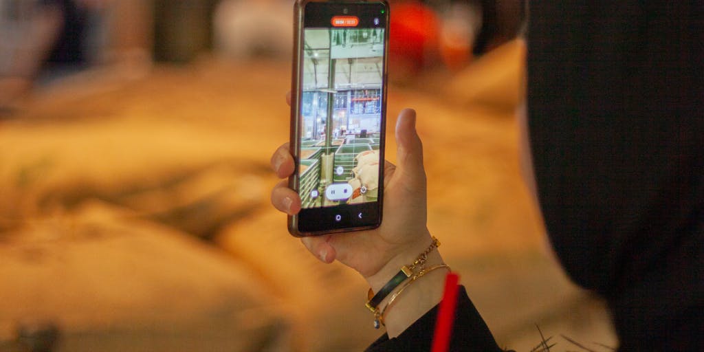 A close-up shot of a hand holding a smartphone displaying the 'Apollo Pricing Unlimited' app interface. The background is a blurred image of a bustling coffee shop, illustrating the convenience and accessibility of the service in everyday life.