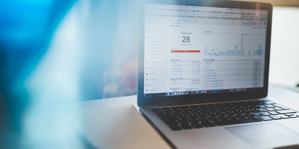 A close-up shot of a laptop screen displaying a Google AdWords dashboard with colorful graphs and metrics. In the background, a person is analyzing data with a notepad and pen, emphasizing the analytical side of advertising.